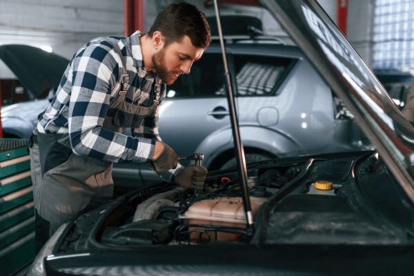 Repairing the electronics of the car under the hood. Man in uniform is working in the auto salon